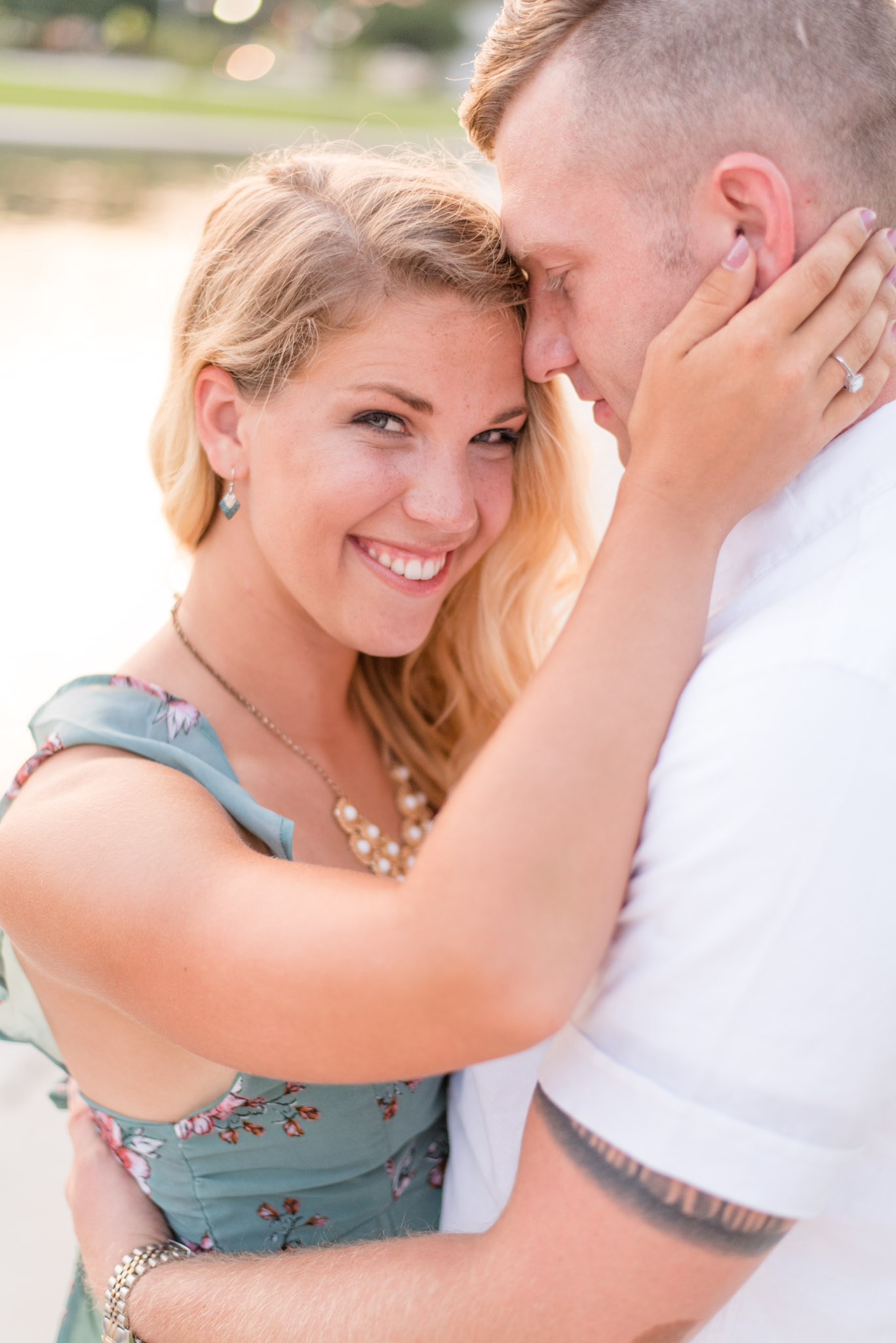 Bride smiles at camera in front of lake during engagement pictures.