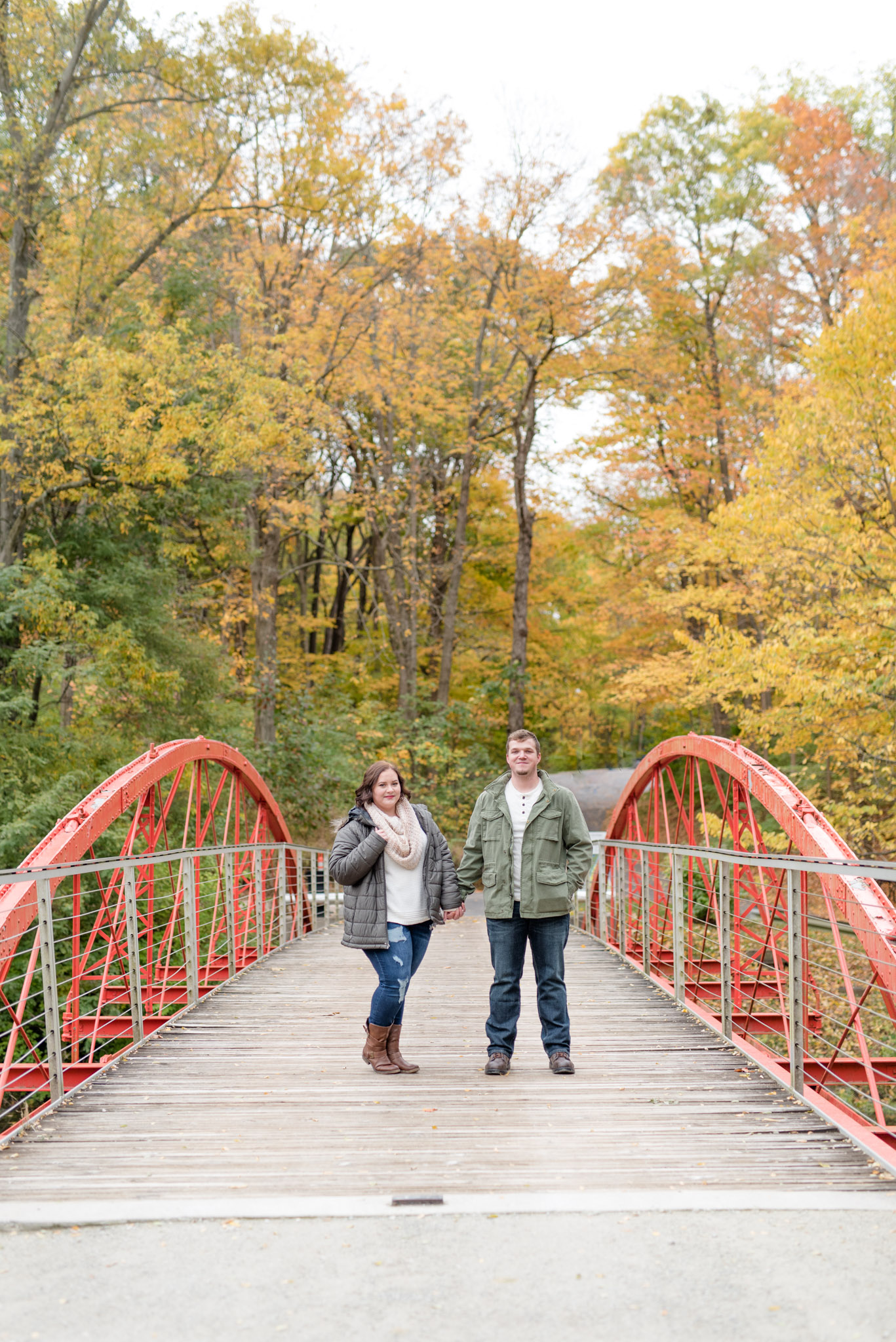 Engaged couple stands on red bridge during fall.
