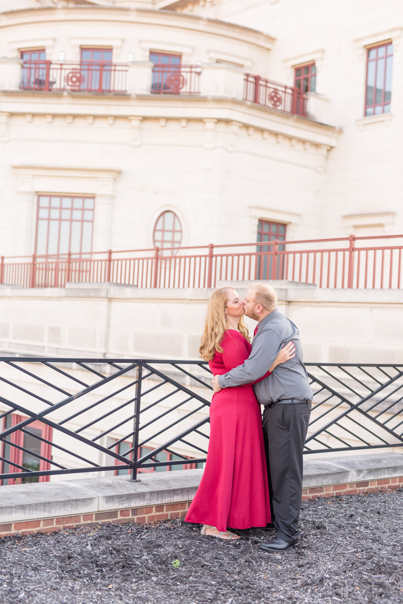 Engaged man and woman kiss in front of white building.