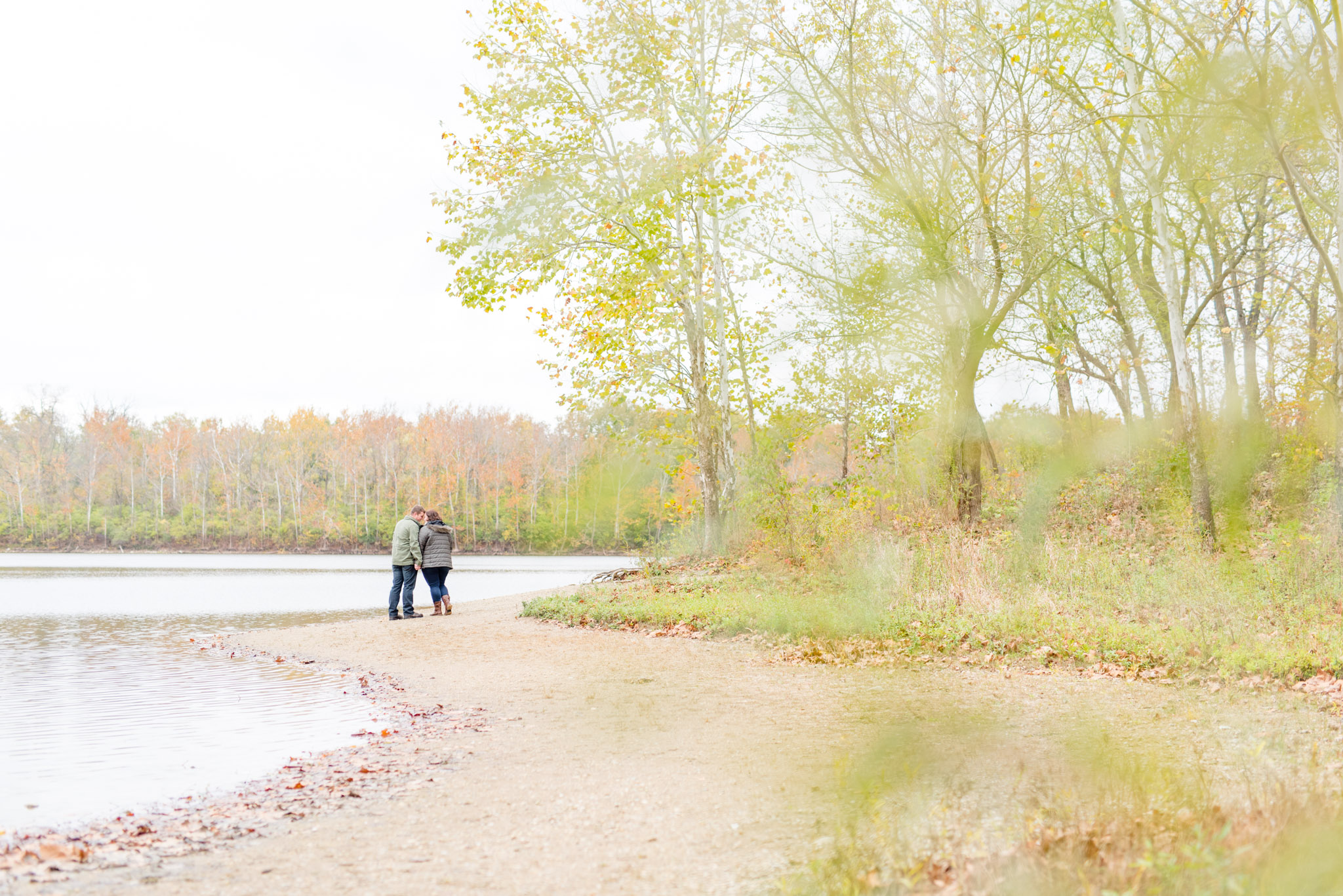 Engaged couple stands on riverbank next to trees.