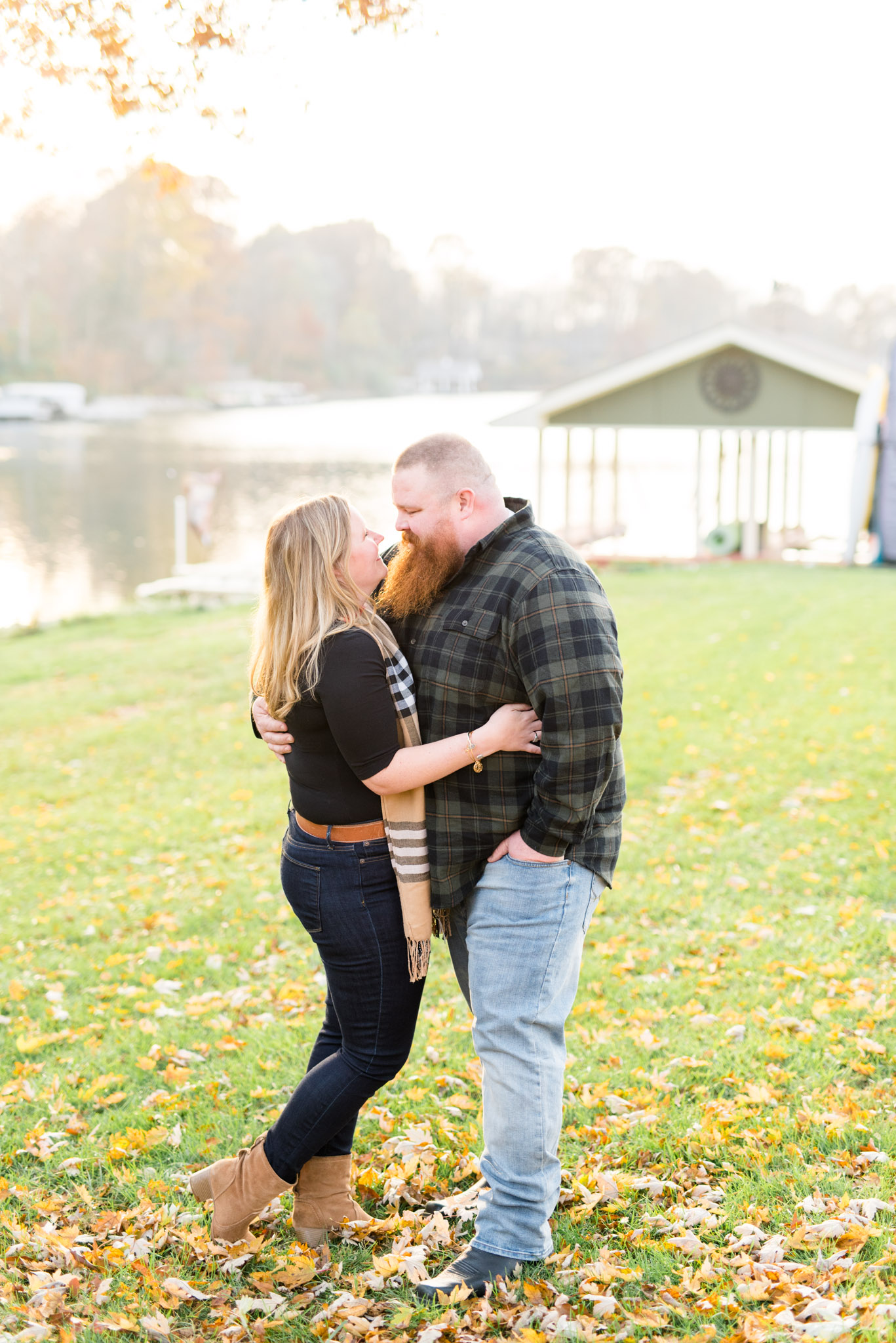 Engaged bride and groom smile at each other near lake during fall.