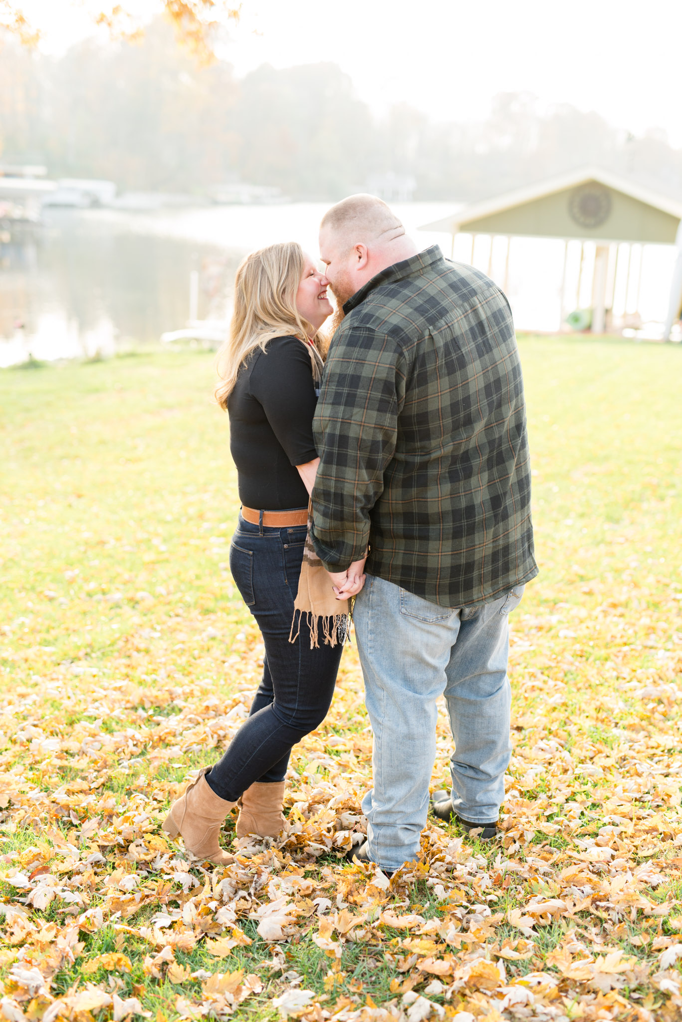 Engaged couple laughs next to lake during fall.