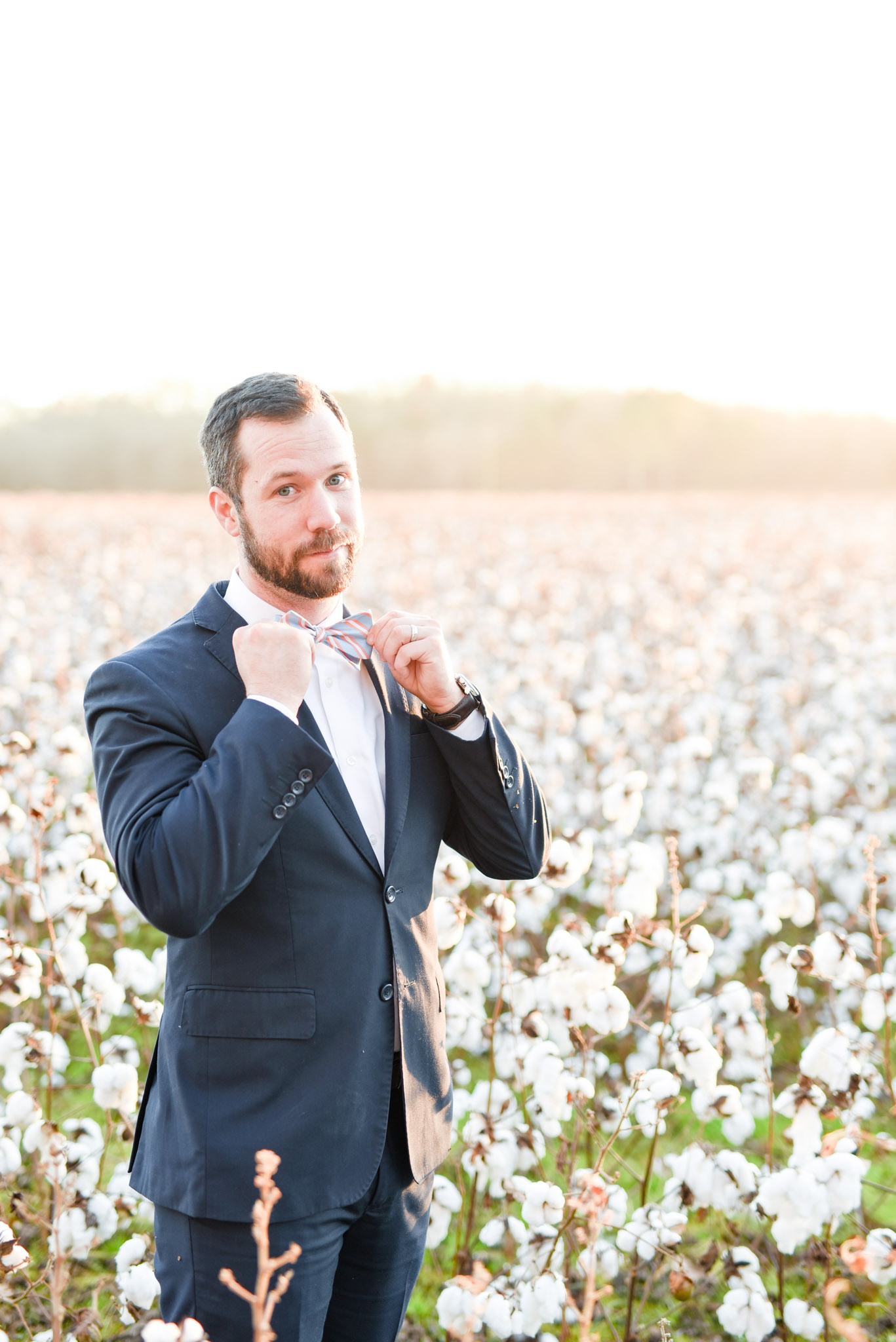 Man straightens bow tie in cotton field at sunset.