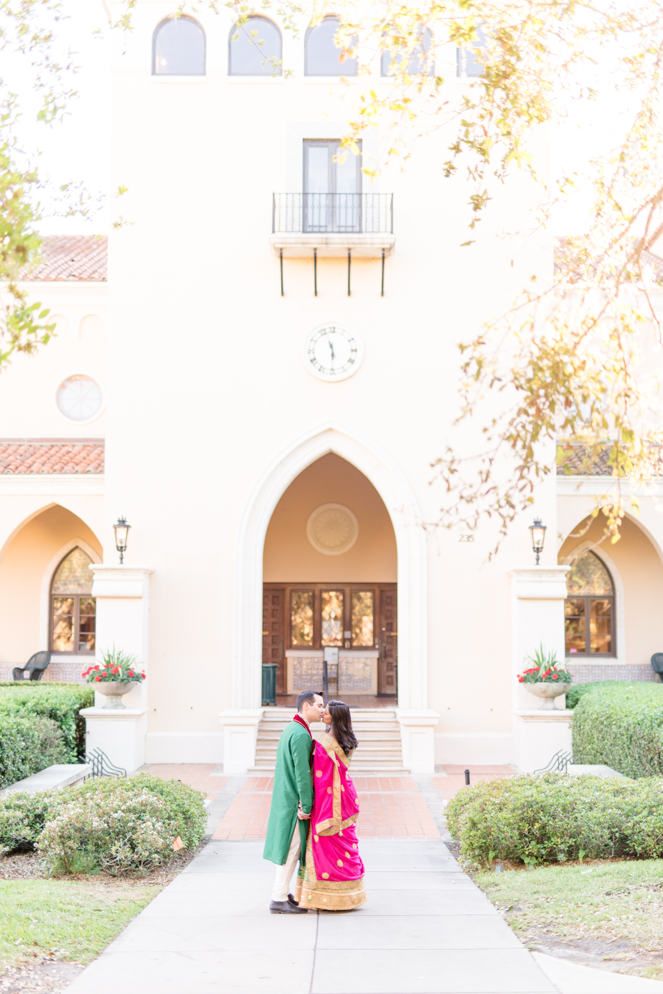 Engaged couple kiss in front of white building.