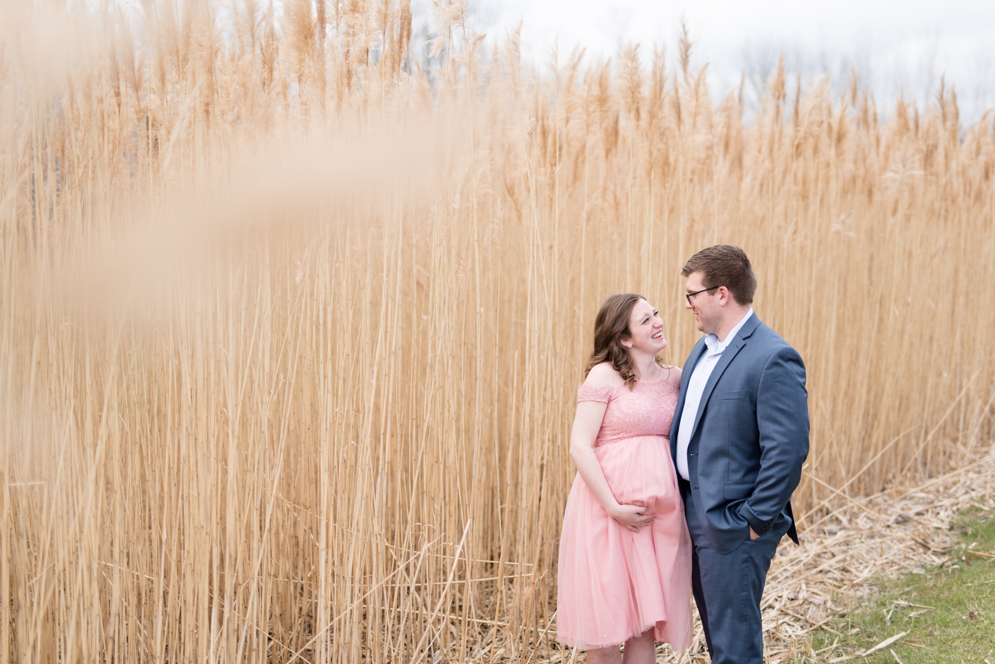 expecting parents laugh next to wheat field.