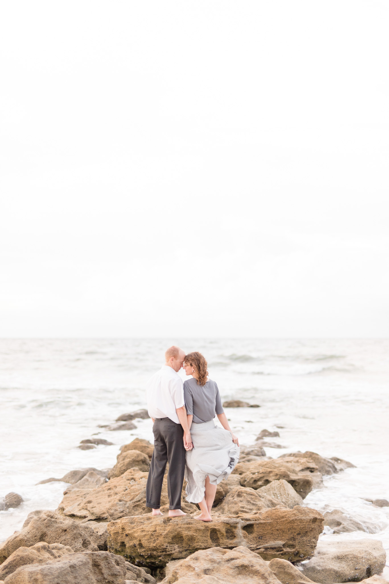 Couple stands on rocks by sea.