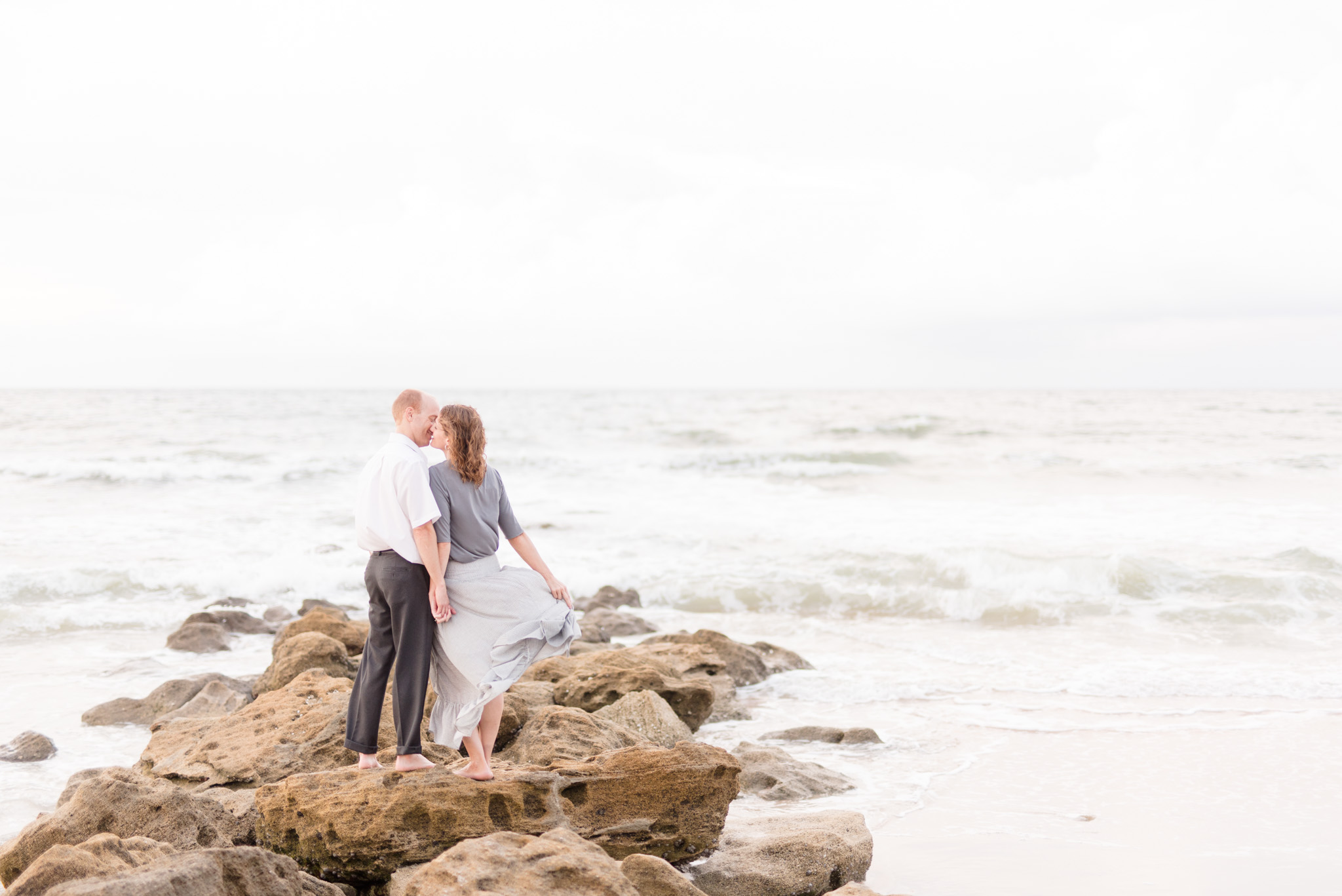 Couple kisses on beach rocks at sunset.