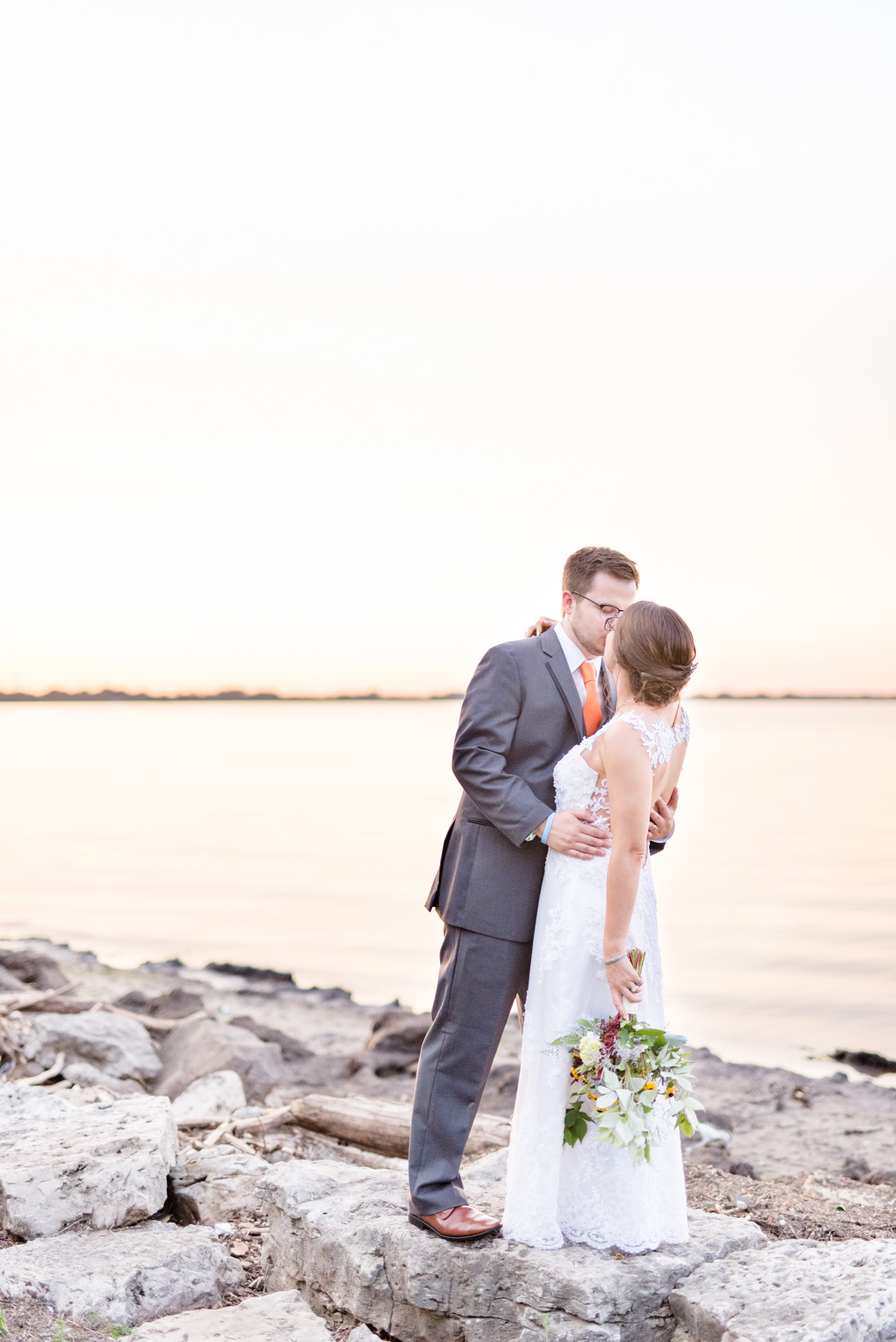 Bride and groom kiss by lake at sunset.