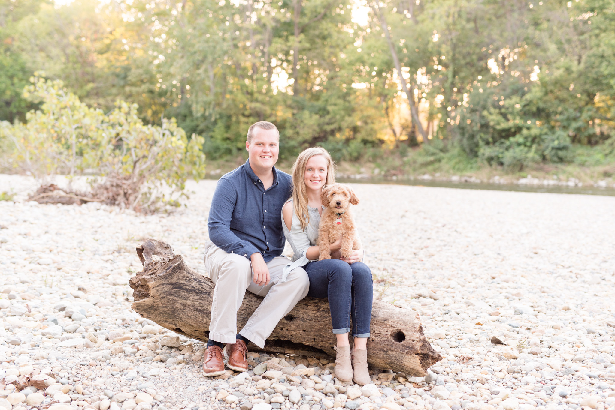 Couple and goldendoodle puppy sits on log.