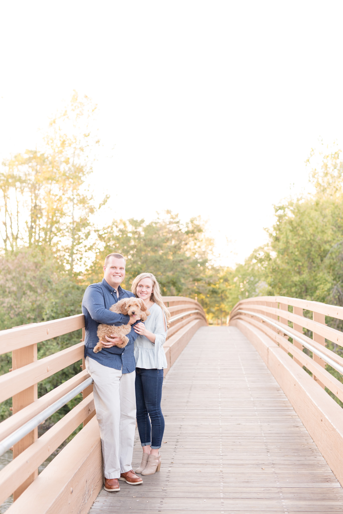 Couple smiles at camera while holding puppy on bridge.