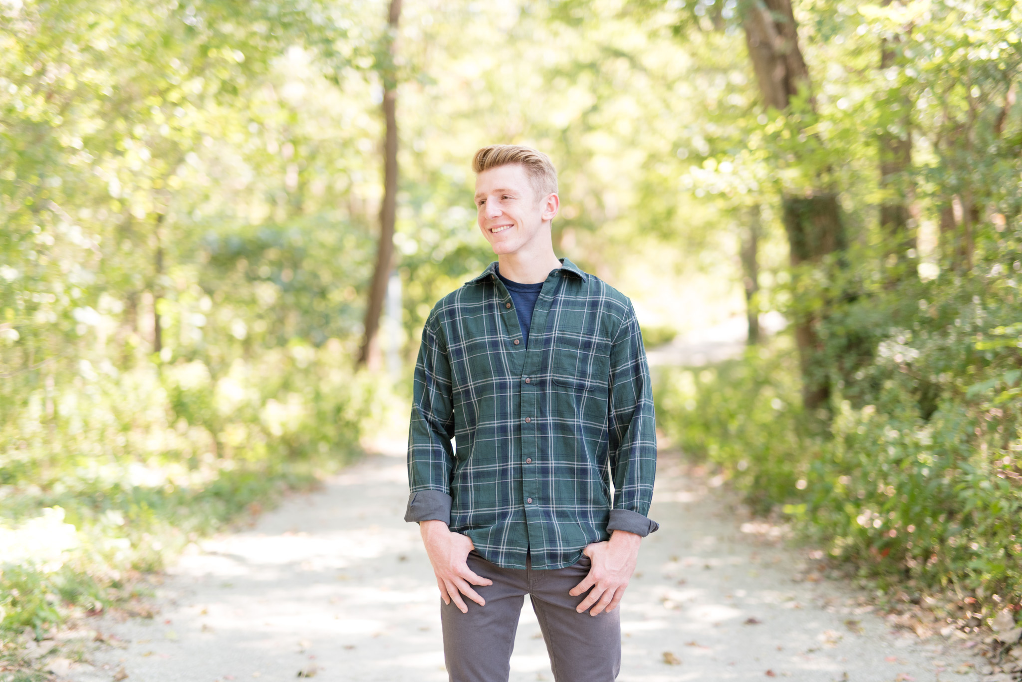 Senior guy wears flannel and stands in forest.