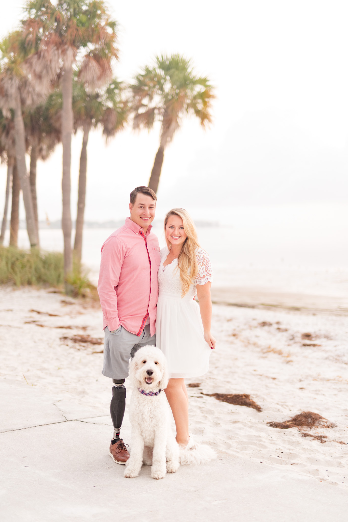 Couple stands on beach with goldendoodle.