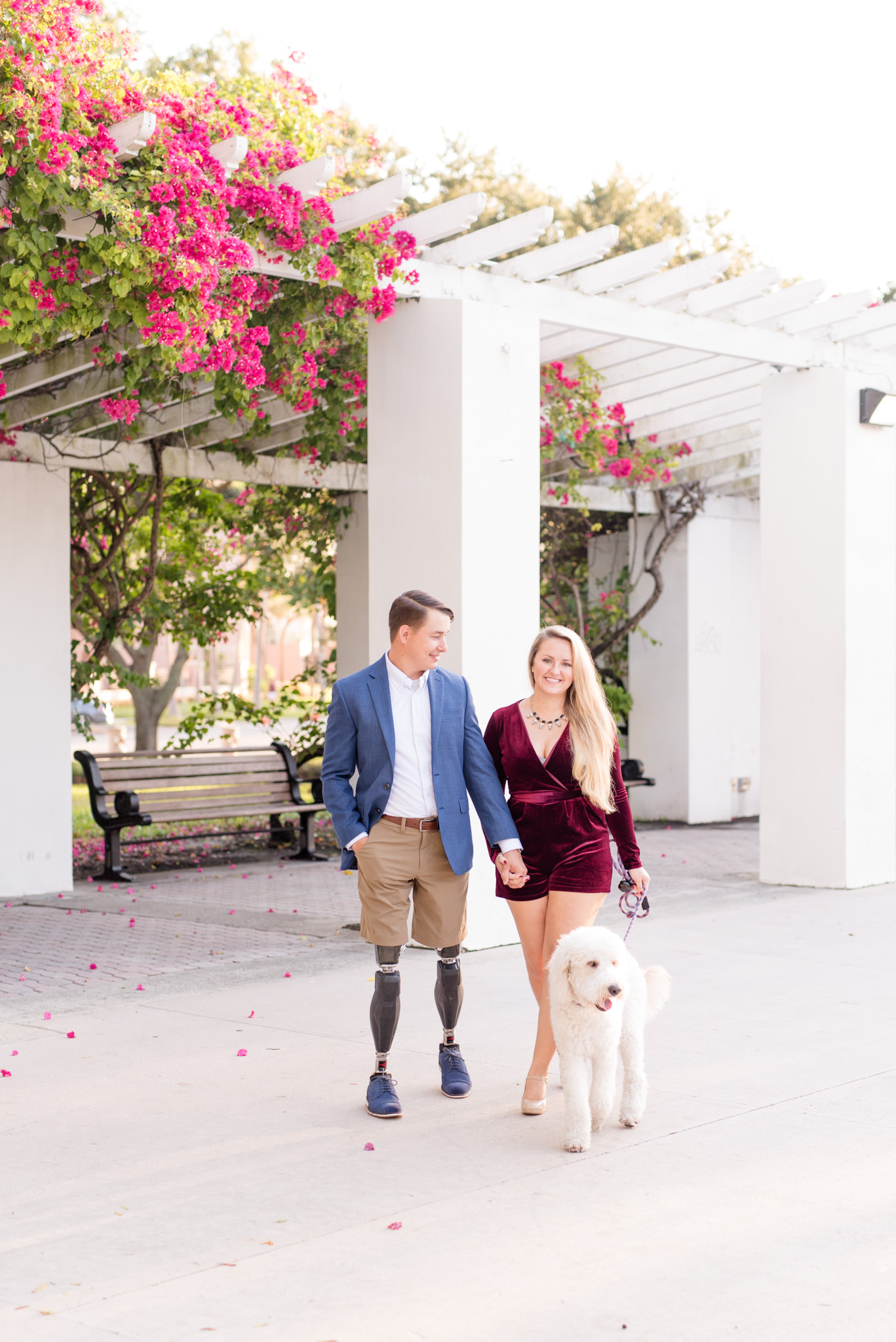 Couple walks with dog in front of flower pergola.