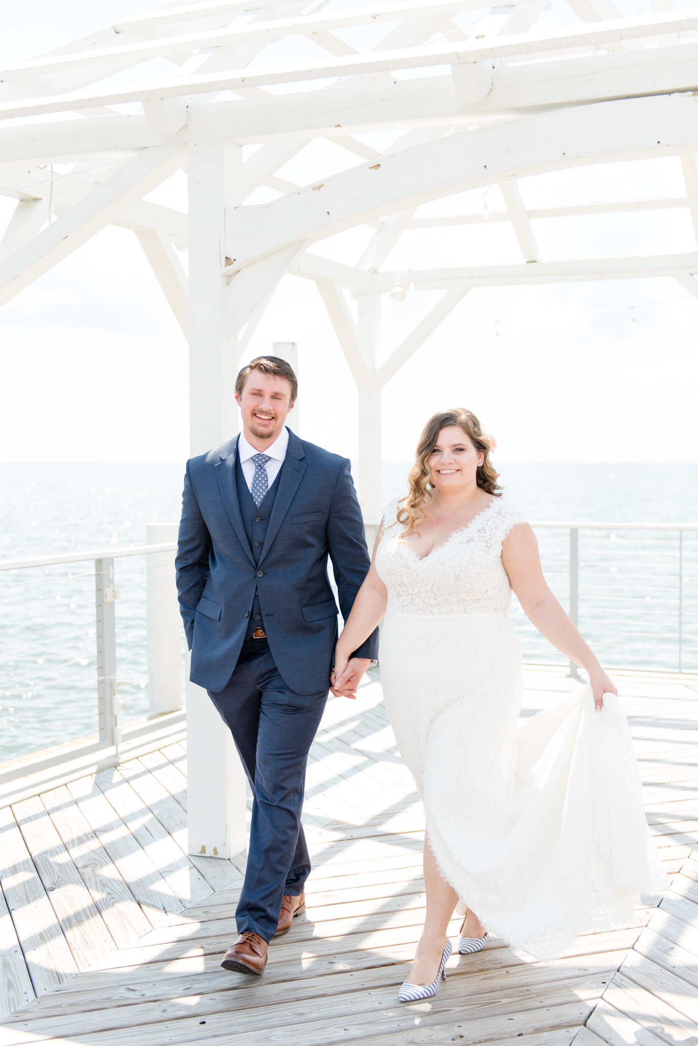 Bride and groom walk on waterfront pier.