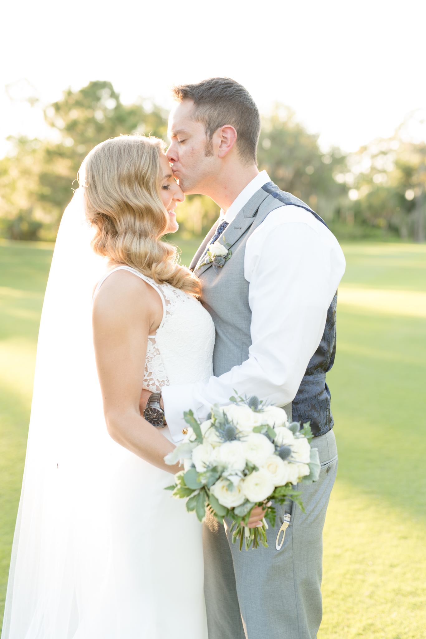 Groom kisses bride on forehead in field.