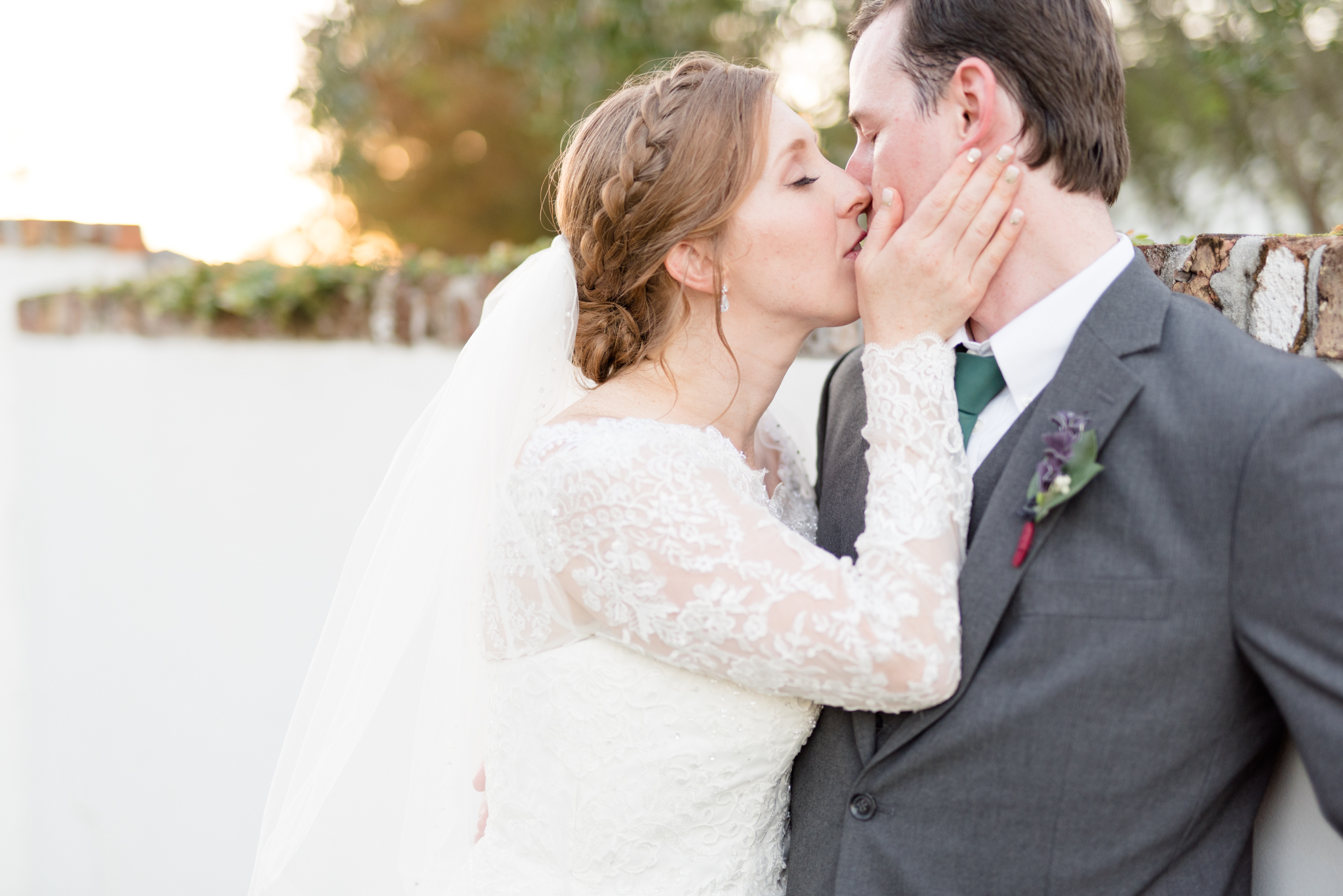 Bride and groom kiss while against white, brick wall.