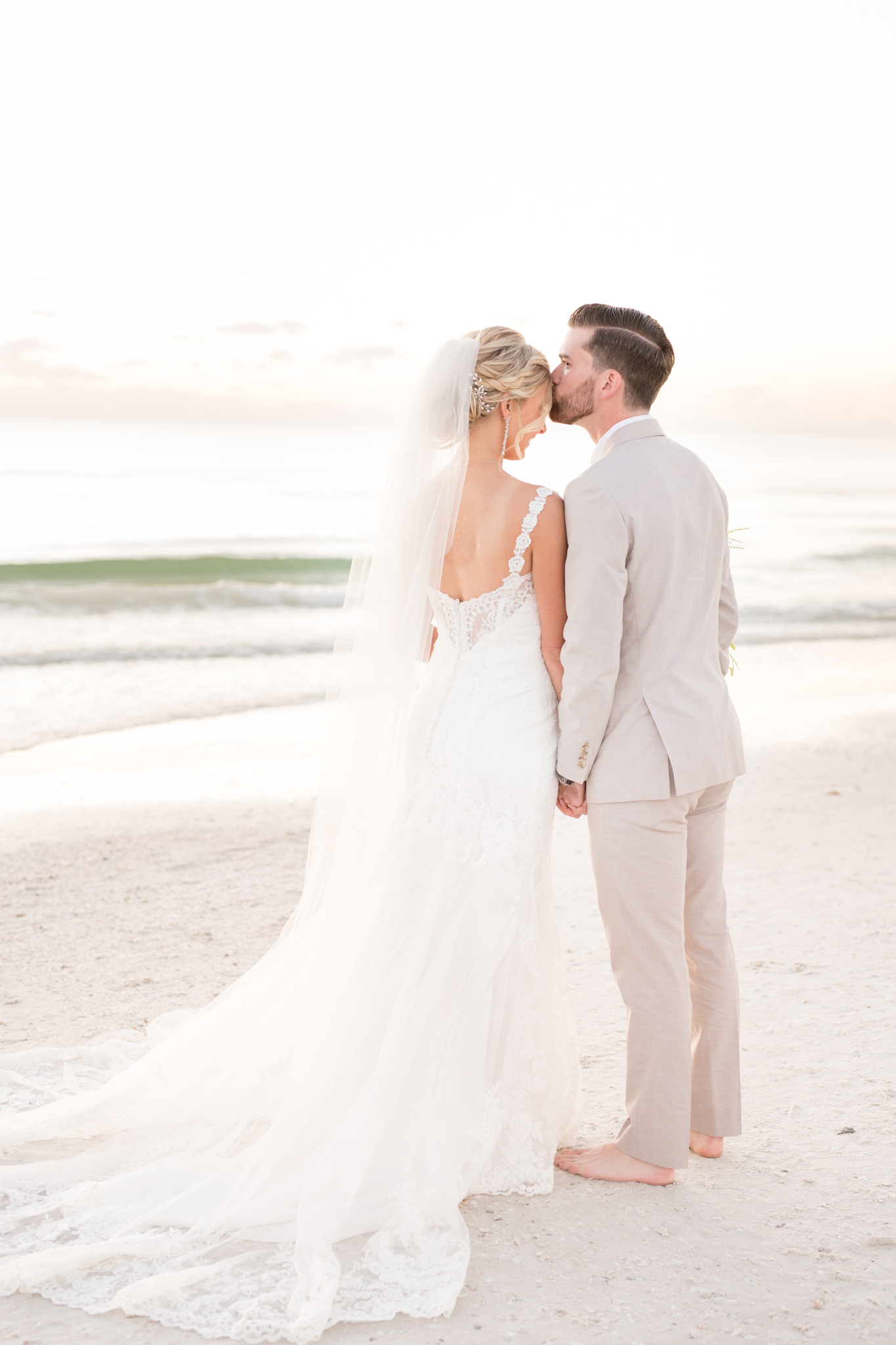 Groom kisses bride on forehead on beach.