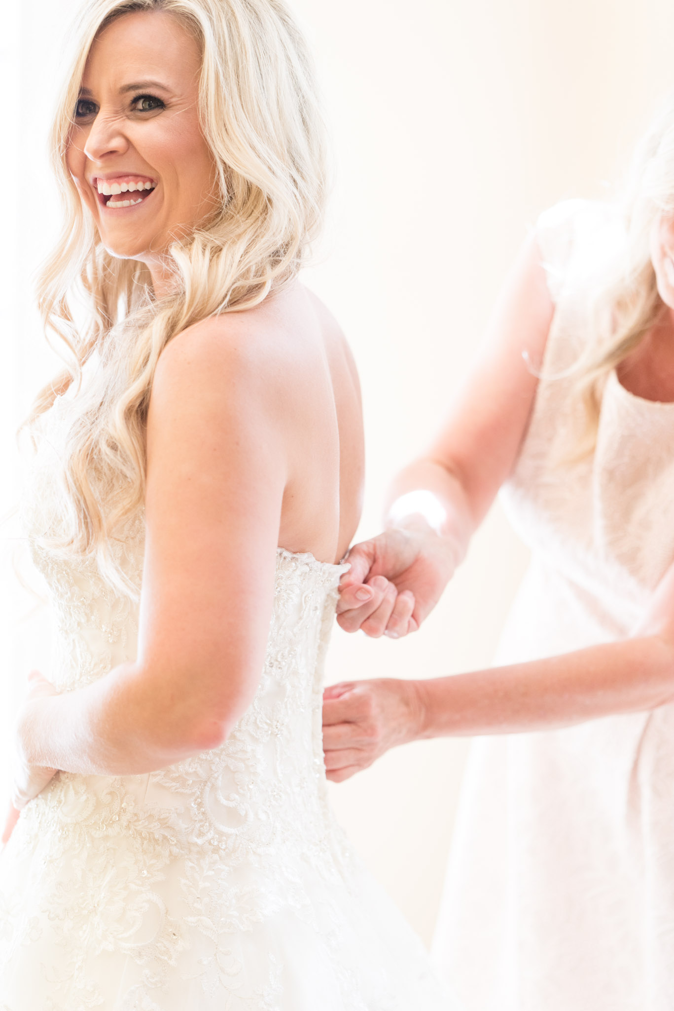 Bride laughs as mother zips dress.