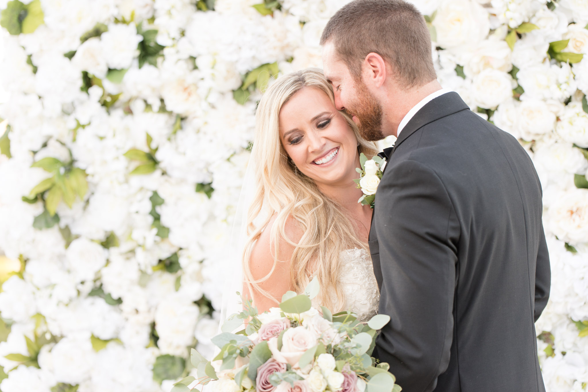 Wedding couple snuggle in front of flower wall.
