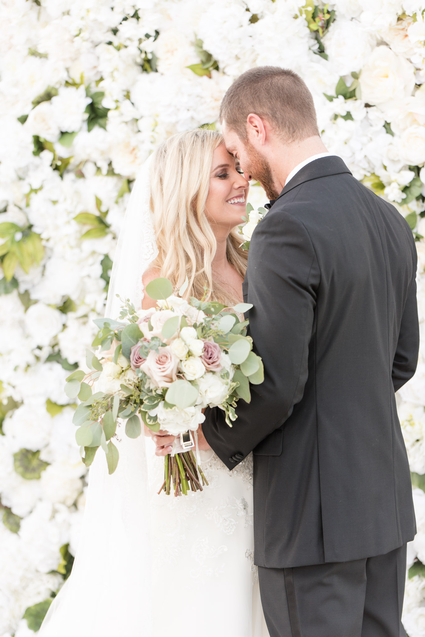 Bride and groom cuddle in front of flower wall.