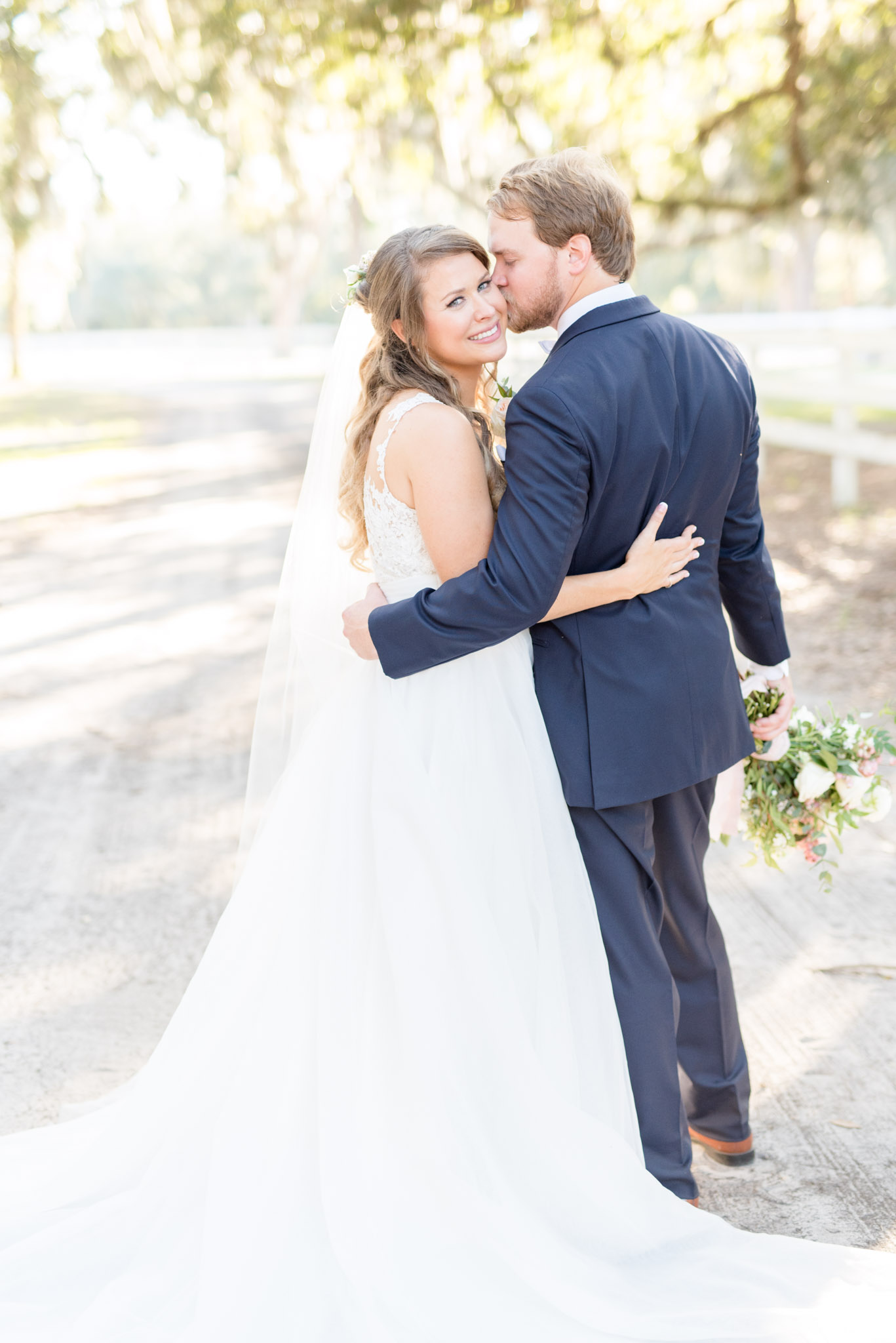 Bride smiles at camera as groom kisses her on cheek.