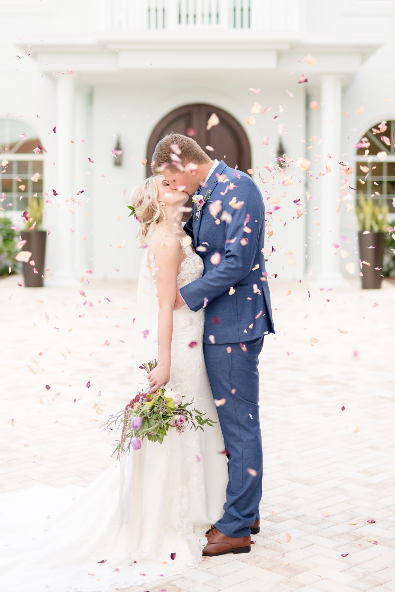 Bride and groom kiss as petals fall around them.