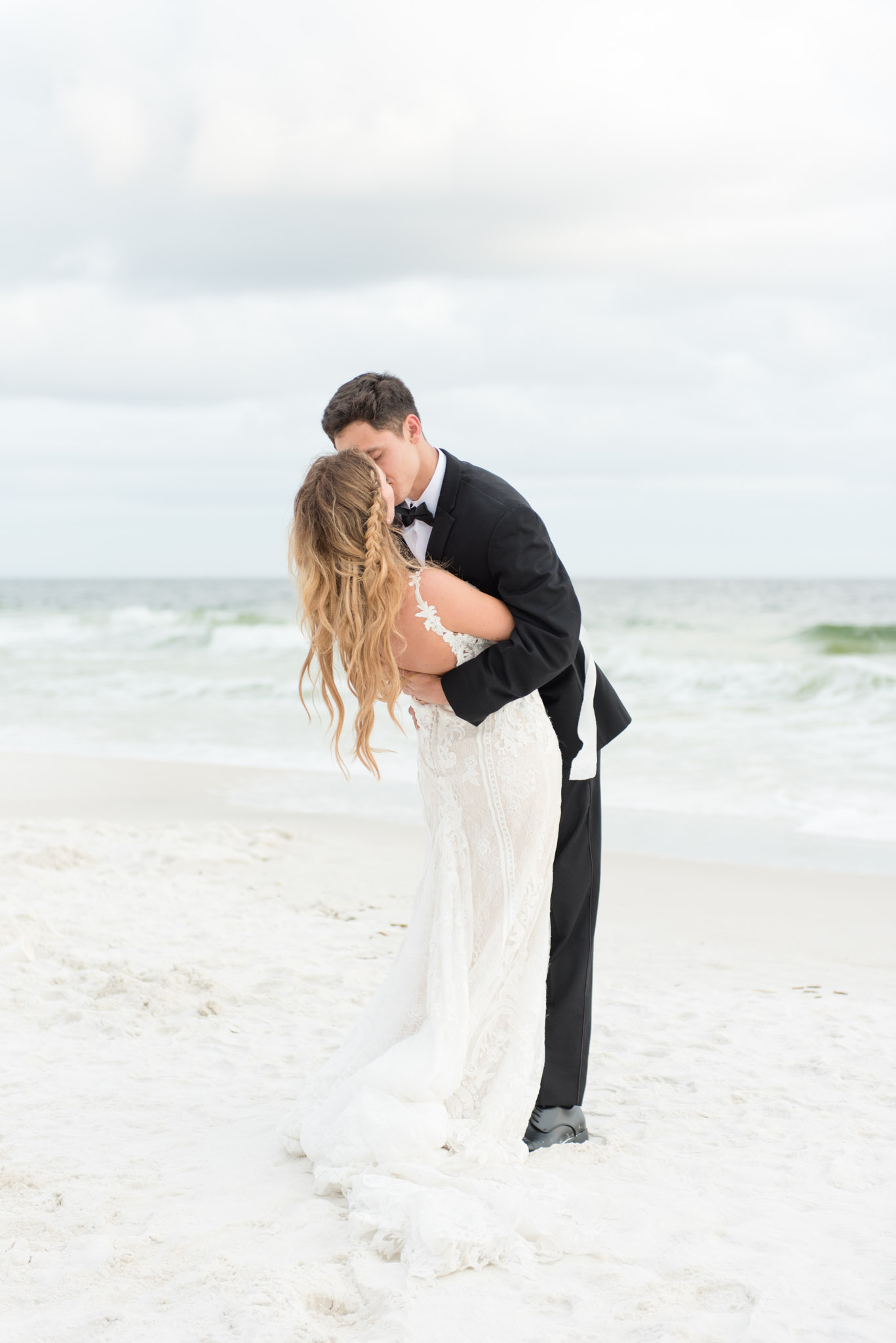 Groom dips bride on beach by ocean.