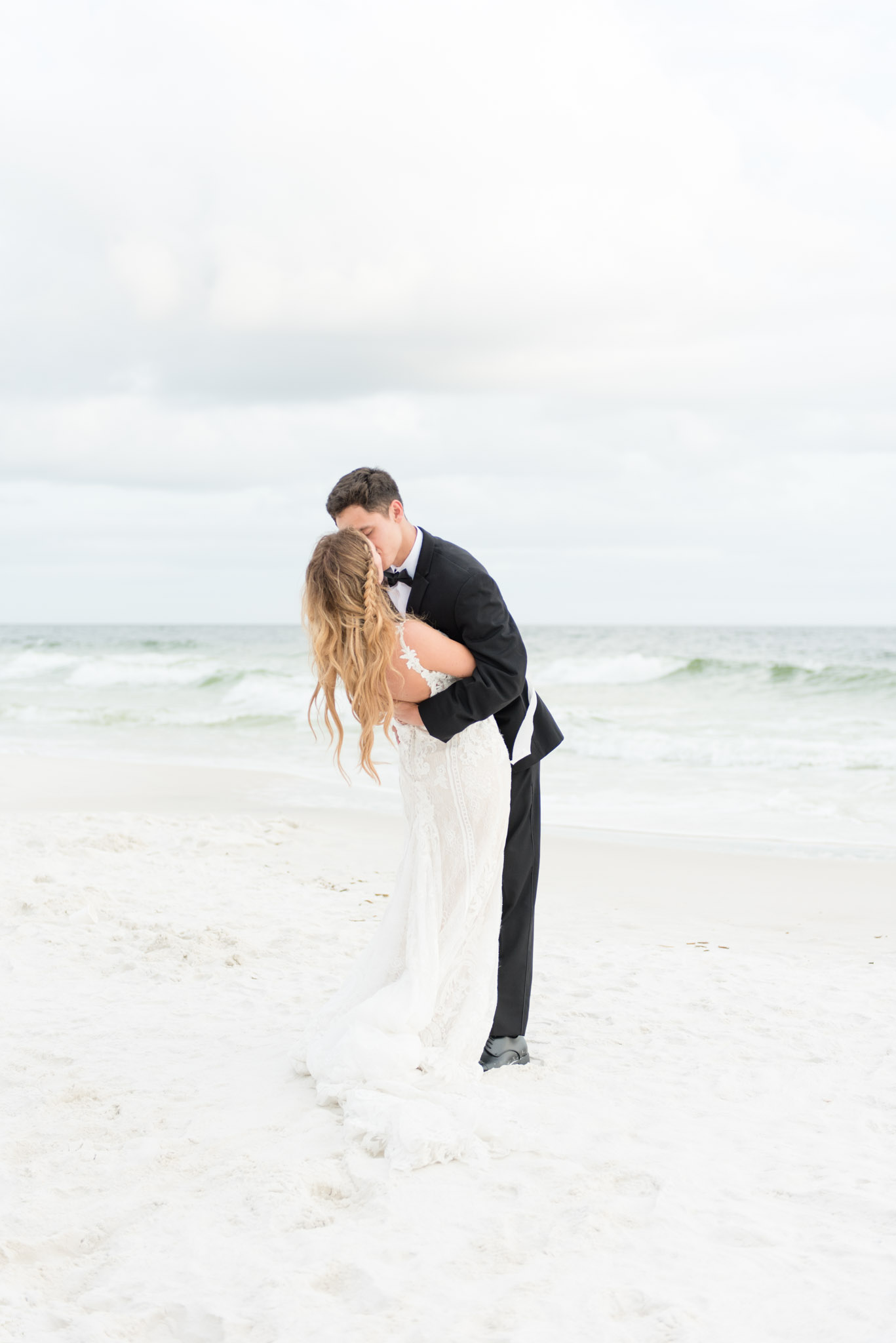 Groom kisses bride on beach by ocean.