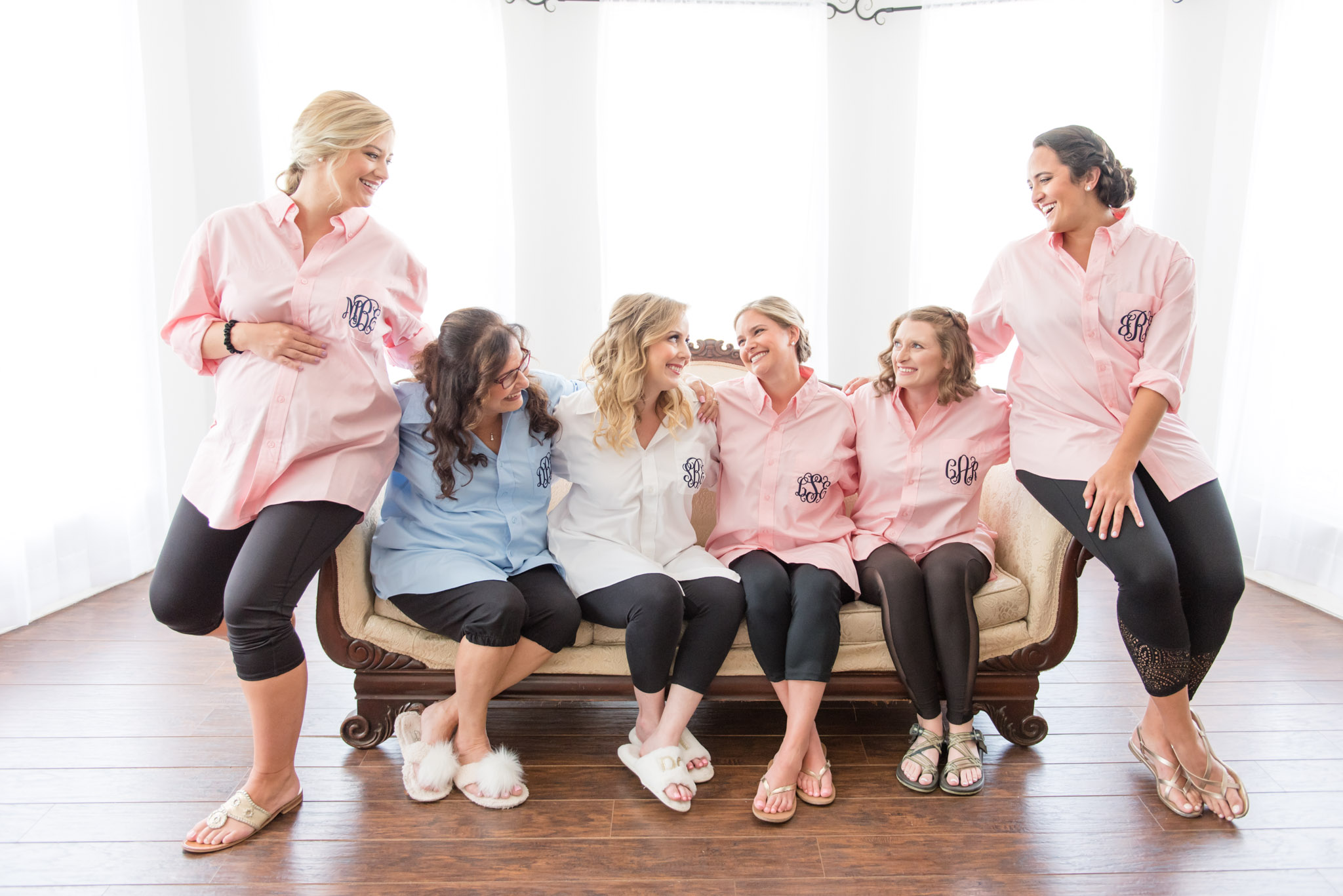 Bride and bridesmaids sit on couch and laugh.