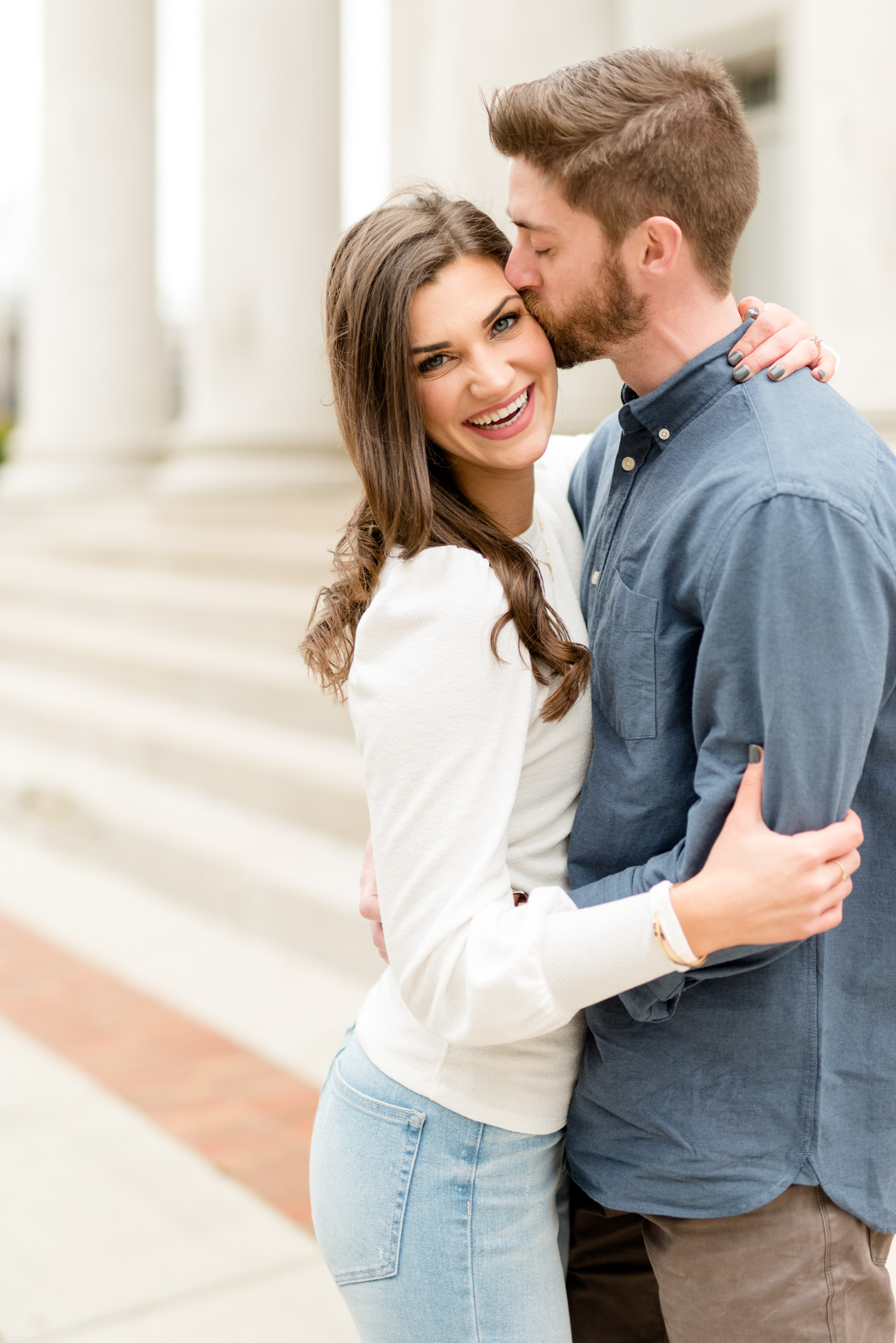 Woman smiles at camera while fiancÃ© kisses her head.