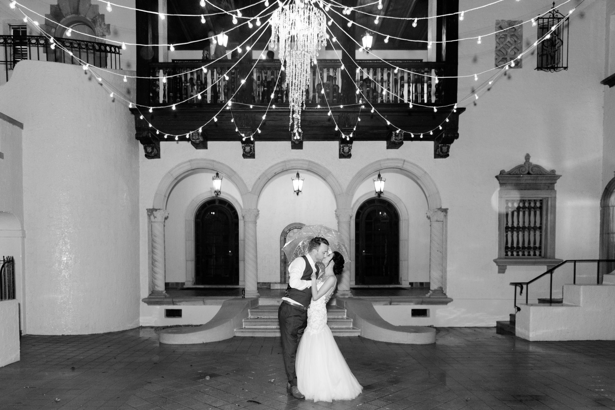 Couple stands under chandelier and kisses.