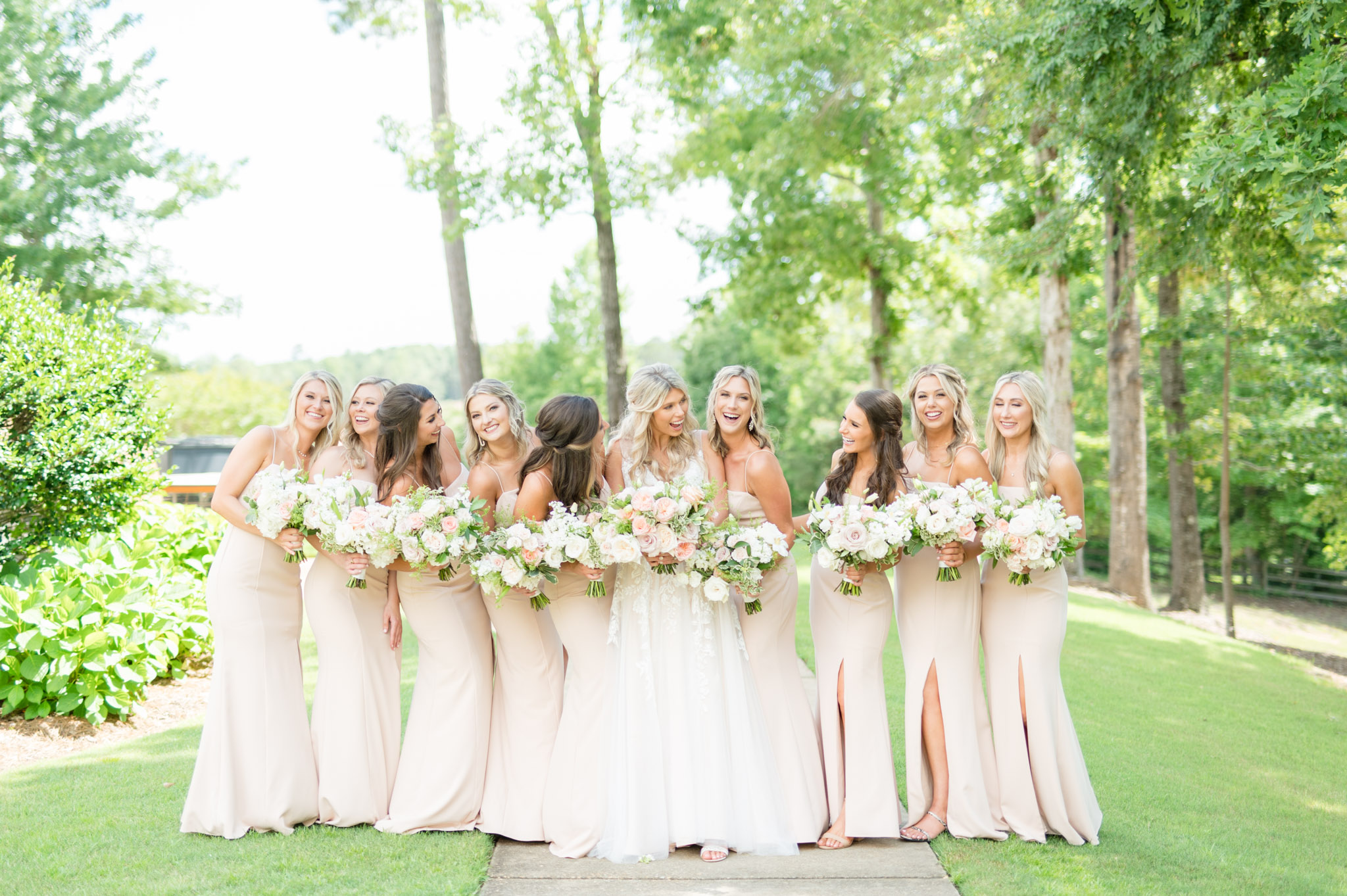 Bride and bridesmaids stand under trees. 