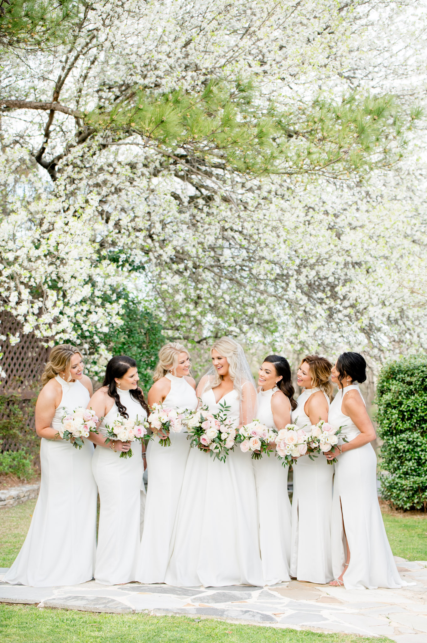 Bride and bridesmaids laugh under blooming dogwood tree.
